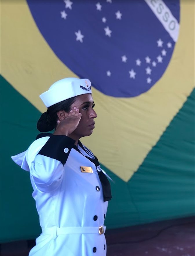 Luana em continência, fardada da Marinha do Brasil, diante da bandeira nacional